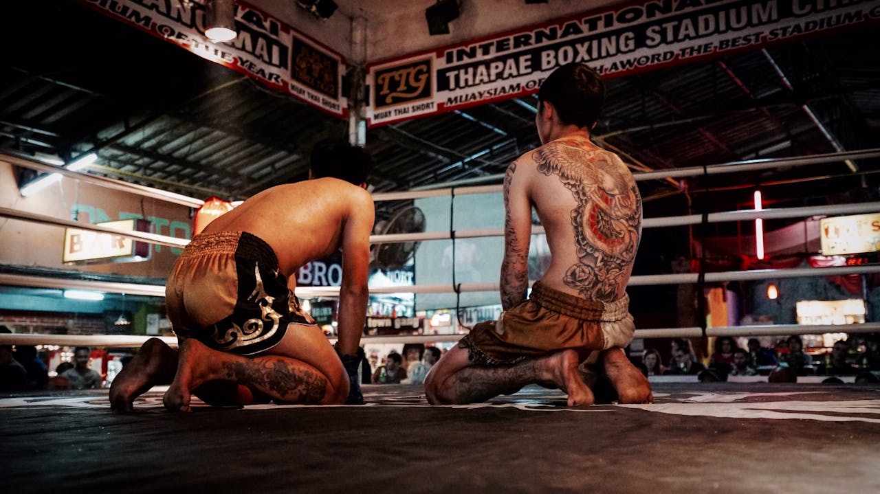 Two tattooed Muay Thai fighters kneeling in a boxing ring at Thapae Boxing Stadium.